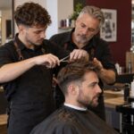 Ein erfahrener Friseurmeister begleitet einen jüngeren Kollegen bei einem Haarschnitt. KI-generiertes Symbolbild.